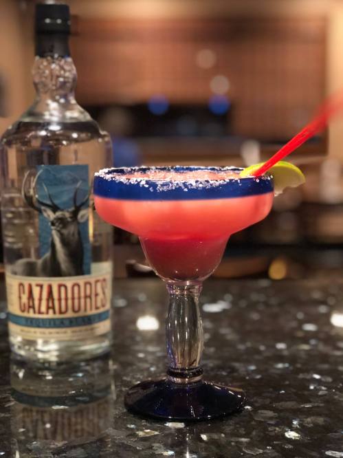 A colorful margarita in a glass with a blue rim, garnished with a lime, next to a bottle of Cazadores tequila.