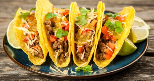 Four tacos filled with beef, tomatoes, cheese, and cilantro, served on a blue plate with lime wedges.