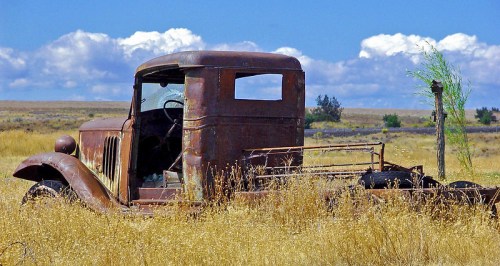 An old, rusty truck sits abandoned in a field of tall grass under a blue sky with fluffy clouds.