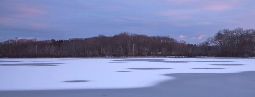 A serene winter landscape featuring a frozen lake with patches of snow and a wooded shoreline under a pastel sky.