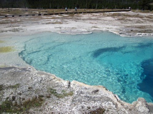 Clear blue geothermal pool surrounded by rocky terrain, with visitors walking on a wooden path in the background.