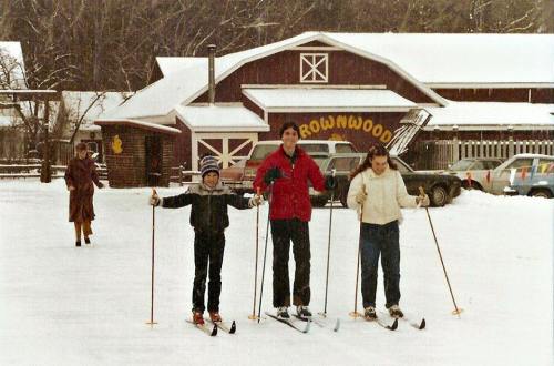 Three people on skis stand in the snow in front of a rustic building, with a car parked nearby and a person walking in the background.