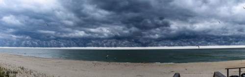 A panoramic view of a beach under a dramatic sky filled with dark clouds, with calm waters and a sandy shore.