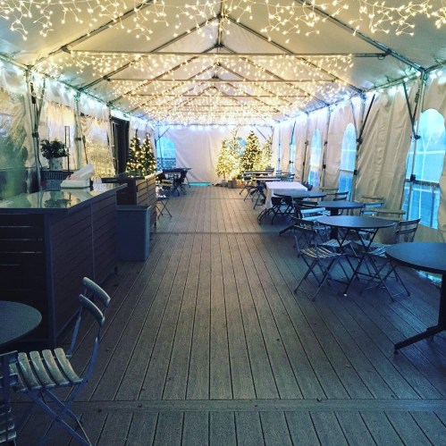 A festive tent decorated with string lights and Christmas trees, featuring tables and chairs arranged along a wooden floor.