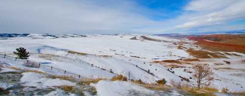 A snowy landscape with rolling hills and a clear blue sky, featuring patches of red earth in the distance.