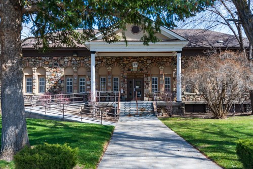 Historic stone building with a front ramp, surrounded by trees and grass, featuring large windows and a welcoming entrance.
