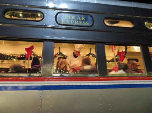 A chef serves treats to passengers inside a decorated Polar Express train, with festive wreaths and lights visible.