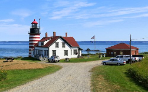 A red and white striped lighthouse beside a white building, with a blue sky and calm water in the background.