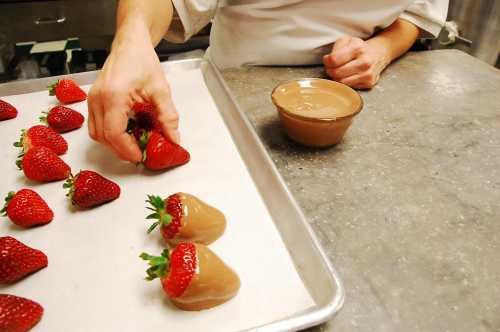 A hand dips strawberries into melted chocolate on a baking tray, with a bowl of chocolate nearby.