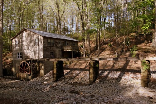A rustic wooden mill with a waterwheel, surrounded by trees and rocky terrain in a serene forest setting.