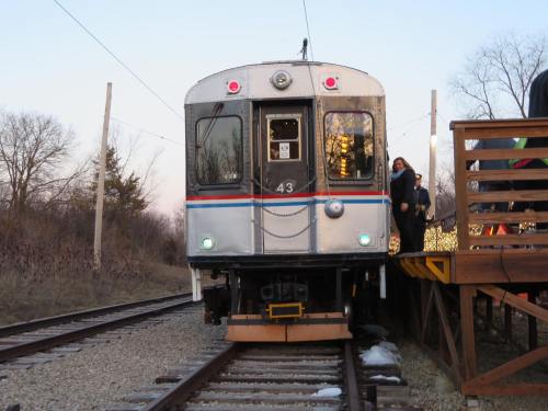A train at a station, with a person stepping off onto a wooden platform, surrounded by trees and a clear sky.