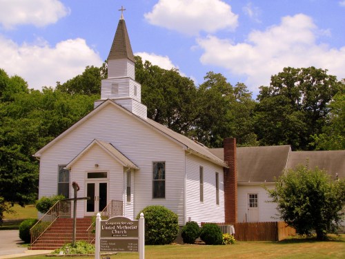 A white church building with a steeple, surrounded by trees and a sign displaying service times.