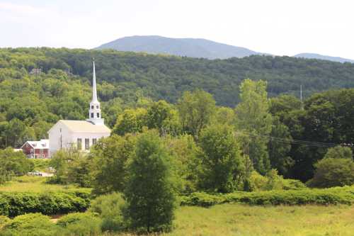 A white church with a tall steeple surrounded by lush greenery and mountains in the background on a sunny day.