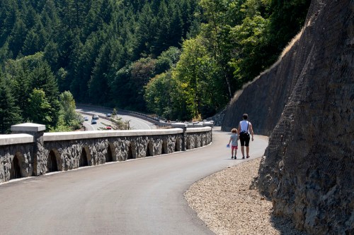 A parent and child walk along a scenic path beside a stone wall, surrounded by lush green trees and a winding road.