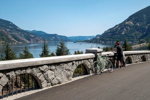 A person leans on a stone railing overlooking a scenic lake surrounded by mountains and trees, with a bicycle nearby.
