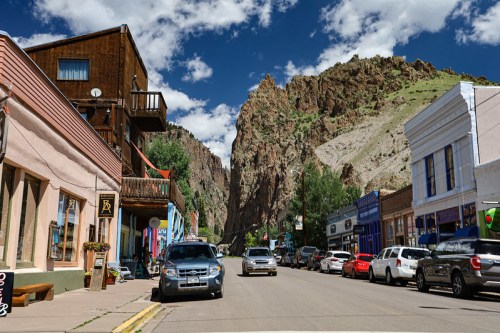 A scenic street lined with colorful buildings and parked cars, framed by towering rocky cliffs under a blue sky.