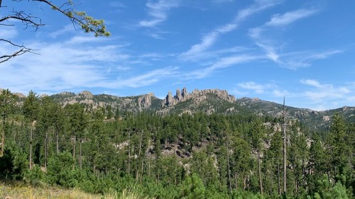 A scenic view of rugged mountains surrounded by lush green forests under a bright blue sky with wispy clouds.