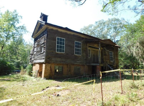 Abandoned, weathered wooden house with broken windows, surrounded by caution tape and overgrown grass.
