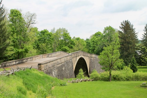 A stone arch bridge spans a green landscape, surrounded by trees and a grassy area under a cloudy sky.