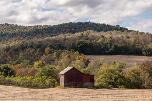 A red barn sits in a field with rolling hills and colorful trees in the background under a cloudy sky.