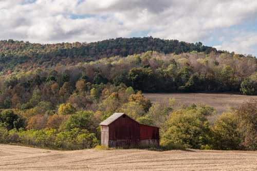 A red barn sits in a field with rolling hills and colorful trees in the background under a cloudy sky.