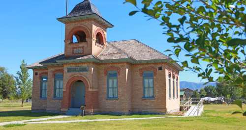 Historic brick building with a tower, surrounded by green grass and trees, under a clear blue sky.