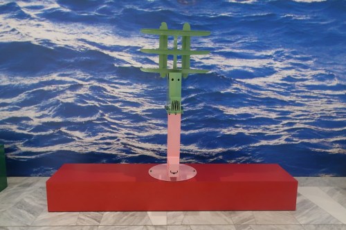 A colorful abstract sculpture stands on a red base against a blue ocean backdrop.