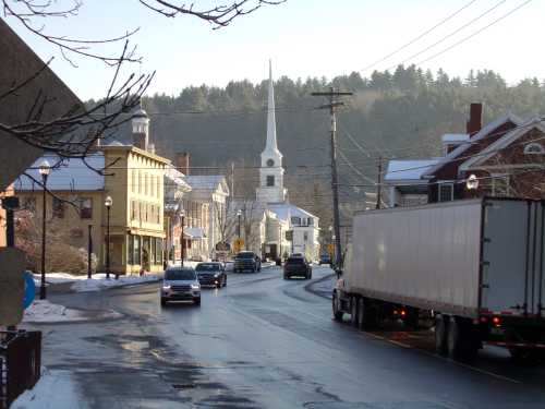 A snowy street scene with cars, a church steeple in the background, and buildings lining the road.