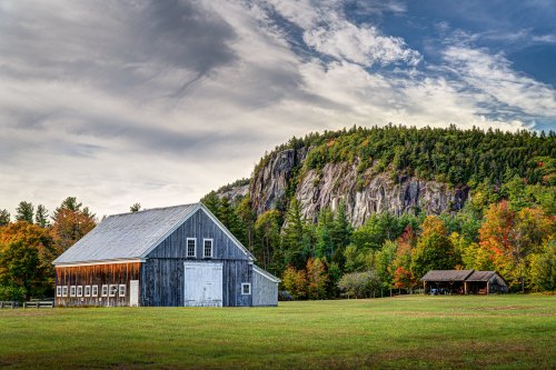 A rustic barn stands in a green field, surrounded by colorful autumn trees and a rocky hillside under a cloudy sky.