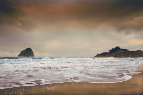 A serene beach scene with gentle waves, rocky formations, and a moody sky at dusk.