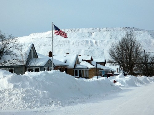 A snowy street with houses and an American flag, set against a backdrop of a snow-covered hill.