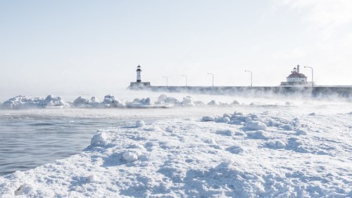 A snowy landscape with a lighthouse and a building in the distance, surrounded by mist rising from the water.