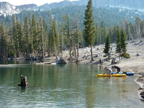 Two people kayak on a calm lake surrounded by trees and mountains, with a sandy shore and rocky landscape.