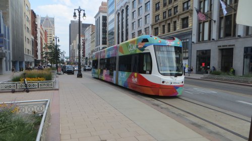 A colorful streetcar travels along a city street, surrounded by buildings and greenery.