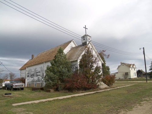 An old, weathered church with a cross on top, surrounded by overgrown shrubs and a cloudy sky.