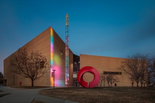 Modern building with colorful lighting and a large red sculpture in the foreground, set against a twilight sky.