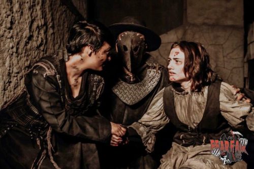 Two characters in tattered clothing interact with a masked figure in a dark, eerie setting.