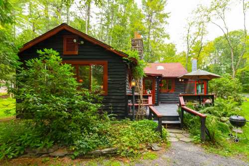 A cozy cabin surrounded by greenery, featuring a stone chimney, wooden deck, and patio umbrella.