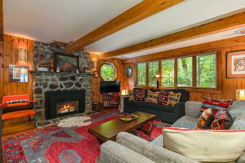 Cozy living room with stone fireplace, wooden beams, large windows, and comfortable seating, surrounded by nature.