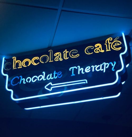 Neon sign for a chocolate café, featuring the text "Chocolate Café" and "Chocolate Therapy" with an arrow.