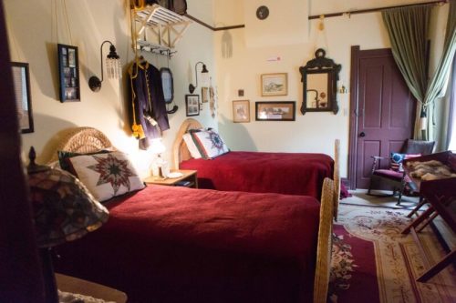 Cozy bedroom with two twin beds, warm red bedding, vintage decor, and a mix of wall art and furnishings.
