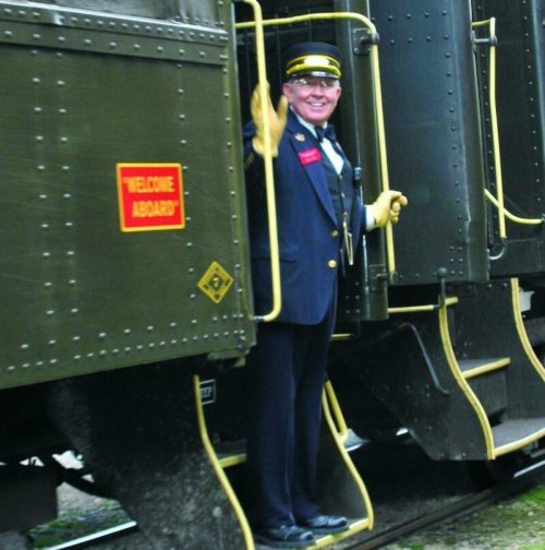 A smiling train conductor waves from the doorway of a train, with a "Welcome Aboard" sign visible nearby.