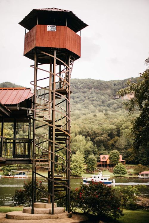 A tall, red observation tower with a spiral staircase overlooks a river and lush green hills in the background.