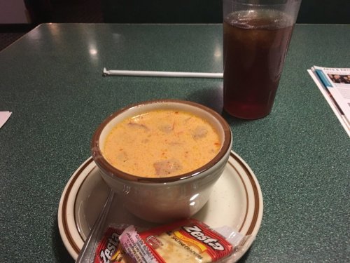A bowl of creamy soup with chunks of meat, served with a packet of crackers and a glass of iced tea on a green table.