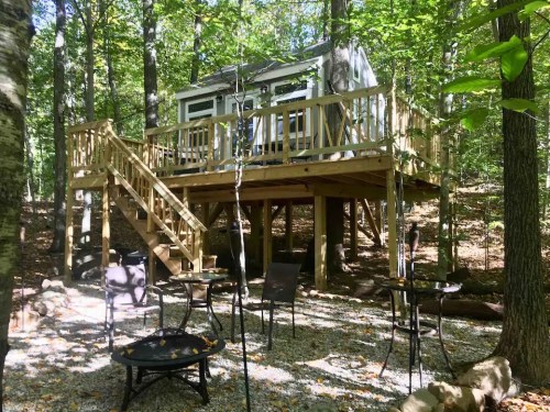 A cozy treehouse on stilts surrounded by trees, with a wooden deck and outdoor seating area below.