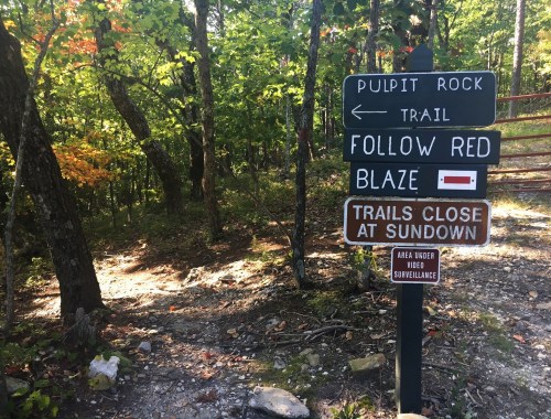 A trail sign in a wooded area, indicating directions to Pulpit Rock and warning that trails close at sundown.