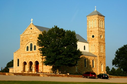 A stone church with a tall bell tower, surrounded by trees and parked cars, under a clear blue sky.