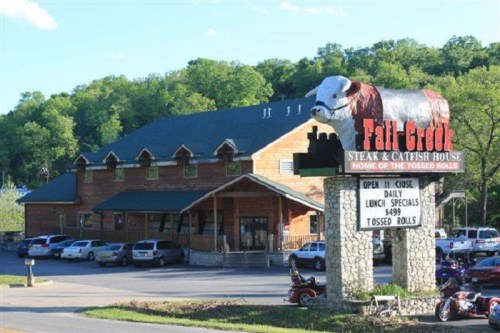 A rustic restaurant with a large cow statue on the roof, featuring a sign for "Fall Creek Steak & Catfish House."