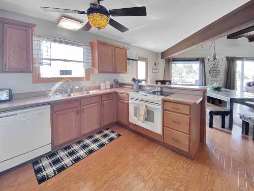 Bright kitchen with wooden cabinets, a ceiling fan, and a checkered rug, featuring a stove and dining area in view.