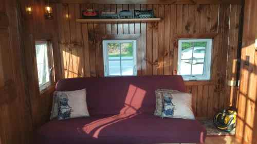 Cozy interior of a wooden room featuring a maroon sofa, decorative pillows, and small windows letting in natural light.